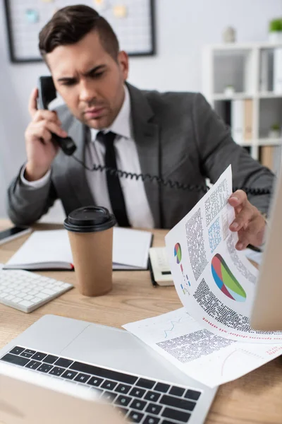 Businessman talking on landline phone while looking at papers with infographics, blurred background — Stock Photo