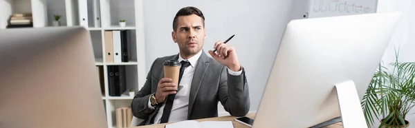 Вдумчивый трейдер держит кофе, чтобы пойти и ручку возле мониторов компьютера, баннер — стоковое фото