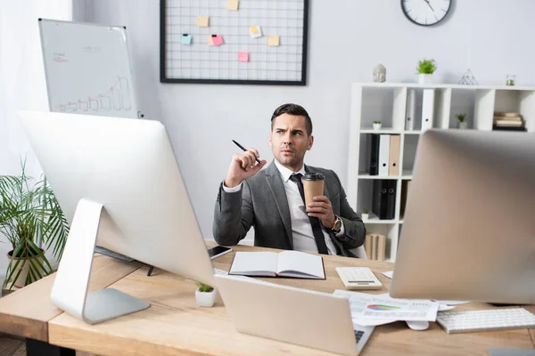 Nachdenklicher Händler mit Coffee to go und Stift in der Nähe von Notizbuch und Monitor am Arbeitsplatz — Stockfoto