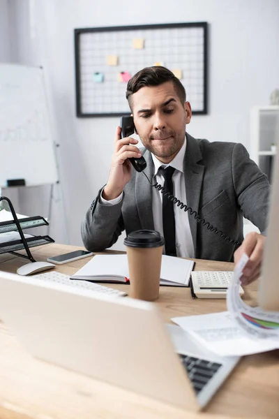 Thoughtful trader talking on landline phone near laptop and coffee to go, blurred foreground — Stock Photo