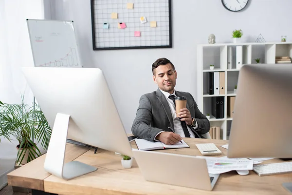 Lächelnder Händler schreibt Notizbuch, während er am Arbeitsplatz mit Coffee to go sitzt — Stockfoto