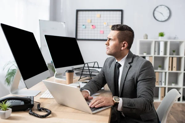 Concentrated trader working on laptop near monitors with blank screen — Stock Photo