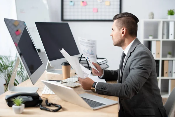 Geschäftsmann hält Infografiken während er am Arbeitsplatz in der Nähe von Monitoren mit leerem Bildschirm sitzt, verschwommener Vordergrund — Stockfoto