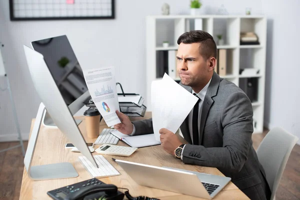 Comerciante reflexivo celebración de infografías cerca de la computadora portátil y monitores en la oficina - foto de stock