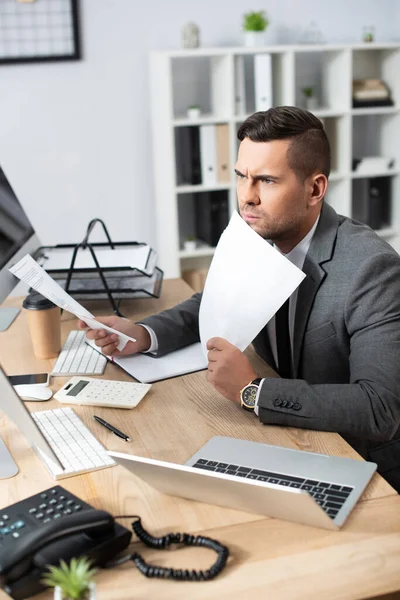 Comerciante serio sosteniendo papeles mientras está sentado en el lugar de trabajo cerca de la computadora portátil, borrosa primer plano - foto de stock