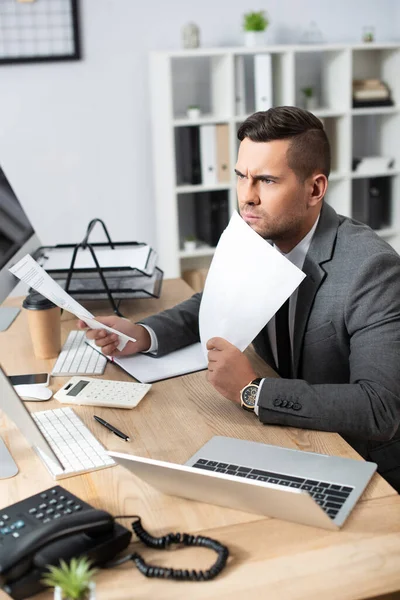 Comerciante grave segurando papéis enquanto sentado no local de trabalho perto do laptop, foreground turvo — Fotografia de Stock