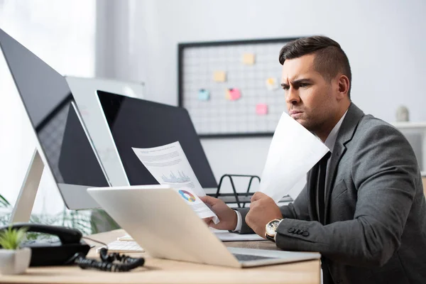 Comerciante serio sosteniendo papeles cerca del ordenador portátil y monitores con pantalla en blanco - foto de stock