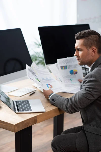 Comerciante reflexivo celebración de infografías cerca de la computadora portátil y monitores con pantalla en blanco - foto de stock