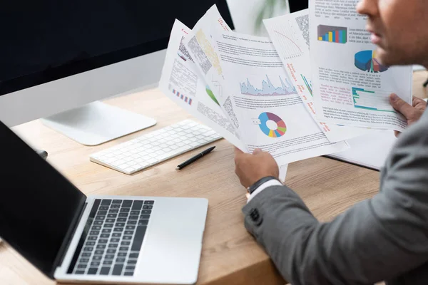 Partial view of trader holding infographics near laptop with blank screen, blurred foreground — Stock Photo