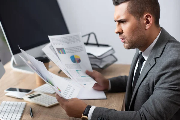 Comerciante serio que trabaja con infografías cerca del monitor de la computadora en la oficina - foto de stock