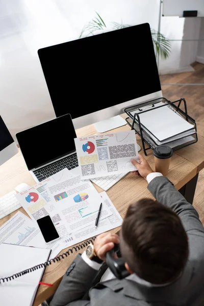 Overhead view of trader talking on telephone near infographics, laptop and monitor, blurred foreground — Stock Photo