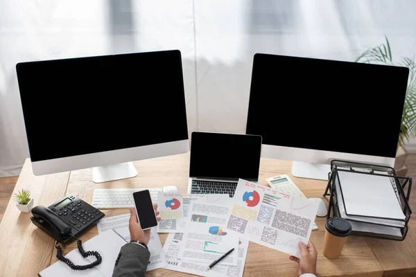 Vista recortada del comerciante que sostiene el teléfono inteligente cerca de infografías, ordenador portátil y monitores con pantalla en blanco - foto de stock