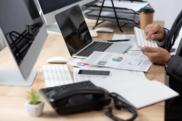 Cropped view of trader typing on keyboard near laptop, monitors, and landline phone on blurred foreground — Stock Photo