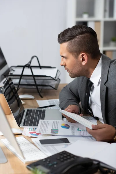 Homme d'affaires concentré regardant ordinateur portable près de papiers avec des graphiques et des graphiques, au premier plan flou — Photo de stock