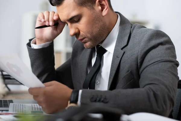 Exhausted trader holding pen and paper while sitting at workplace with closed eyes, blurred foreground — Stock Photo