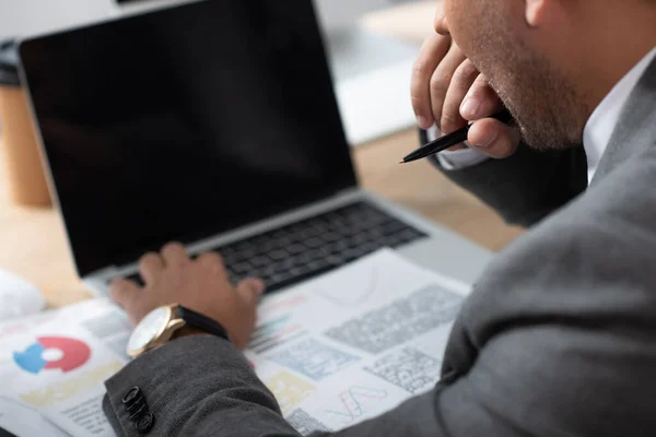 Vista ritagliata del commerciante digitando sul computer portatile vicino a carte con grafici, sfondo sfocato — Foto stock