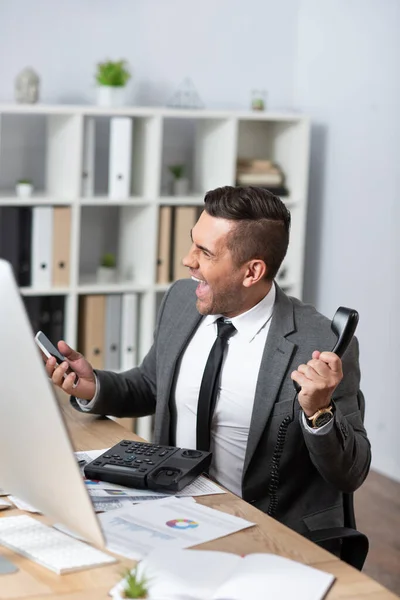 Excited trader screaming while holding handset and smartphone at workplace, blurred foreground — Stock Photo