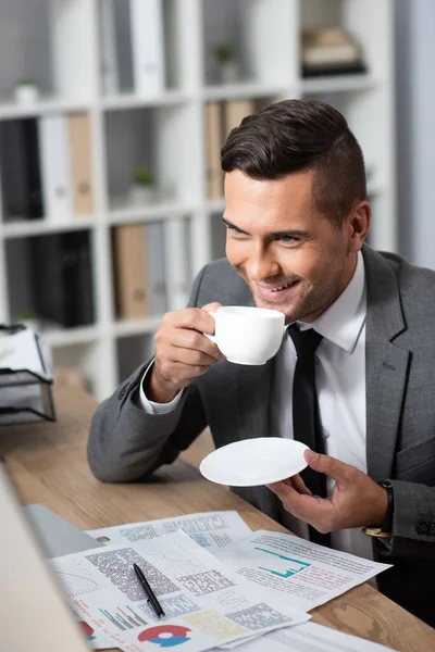 Smiling businessman holding coffee cup at workplace near infographics, blurred foreground — Stock Photo