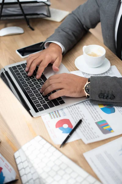 Partial view of trader typing on laptop near coffee cup and papers with infographics — Stock Photo