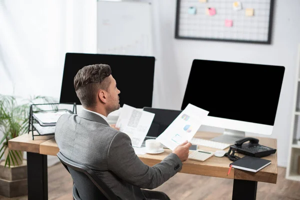 Comerciante segurando infográficos perto de monitores com tela em branco — Fotografia de Stock