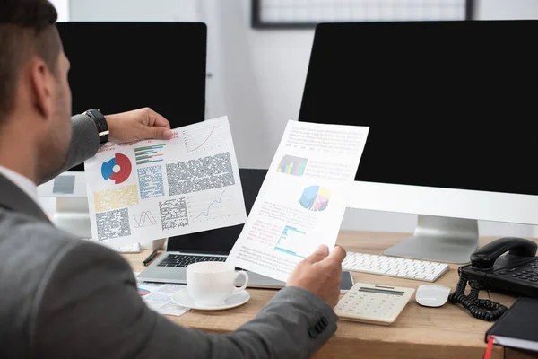 Cropped view of trader looking at papers with charts near computer monitors and coffee cup, blurred foreground — Stock Photo