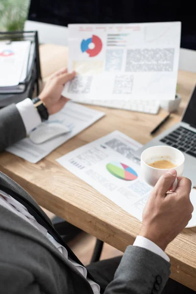 Cropped view of trader holding coffee cup and infographics, blurred background — Stock Photo