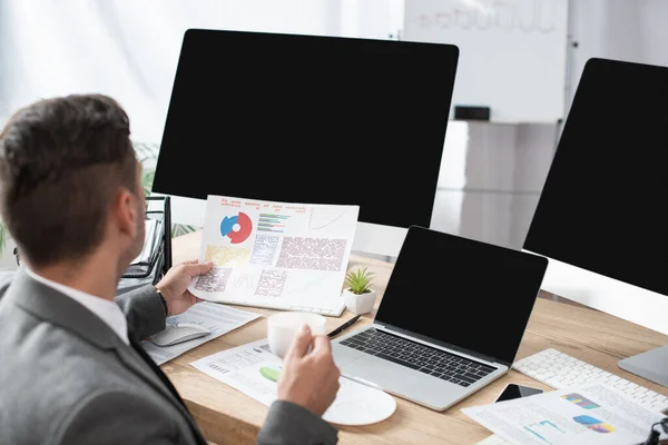 Trader holding infographics near laptop and monitors with blank screen, blurred foreground — Stock Photo