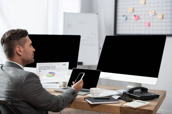 Trader holding smartphone near laptop and computer monitors with blank screen — Stock Photo