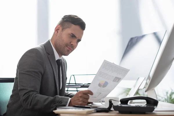 Thoughtful trader looking at paper with infographics near computer monitors — Stock Photo