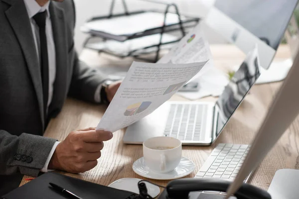 Vista ritagliata del commerciante in possesso di carte con infografica vicino laptop e tazza di caffè — Foto stock