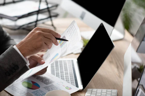 Vista recortada del comerciante apuntando con pluma a las infografías cerca del ordenador portátil con pantalla en blanco — Stock Photo