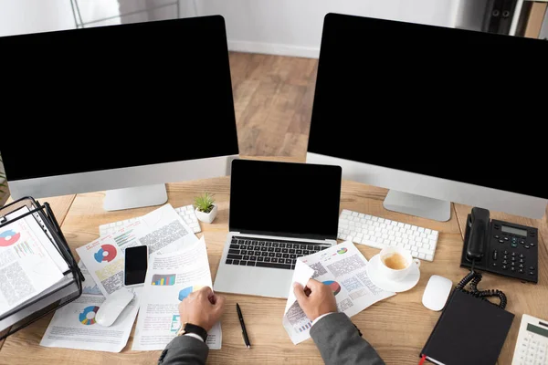 Vista recortada del comerciante cerca de infografías, ordenador portátil y monitores con pantalla en blanco - foto de stock