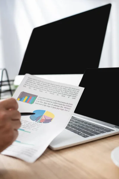 Vista recortada del comerciante apuntando con pluma a las infografías cerca del ordenador portátil y monitor con pantalla en blanco, primer plano borrosa - foto de stock