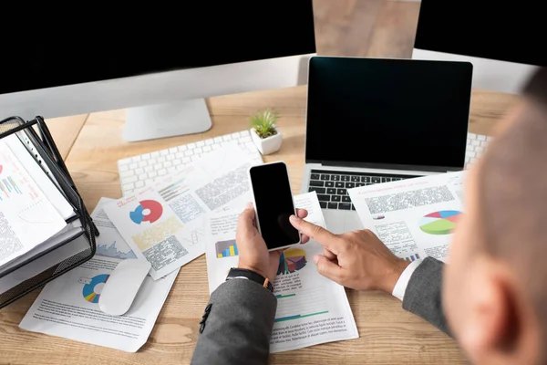 Частичное представление трейдера, указывающего пальцем на смартфон рядом с ноутбуком, мониторы и инфографика, размытый передний план — стоковое фото