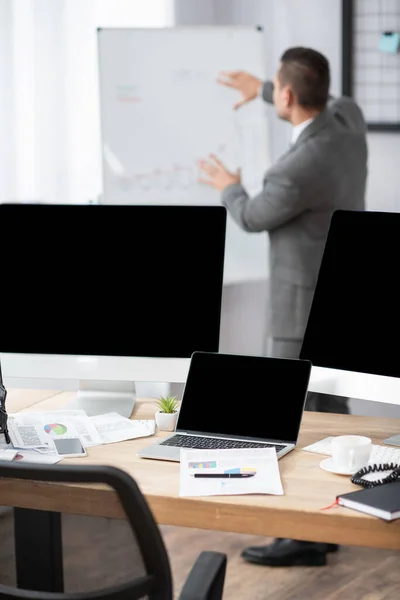 Laptop and computer monitors with blank screen near trader pointing with hands at flipchart on blurred background — Stock Photo