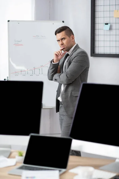 Thoughtful trader standing at flipchart with graphs near monitors with blank screen, blurred foreground — Stock Photo
