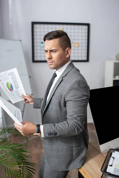 Thoughtful trader in formal wear holding infographics in office — Stock Photo
