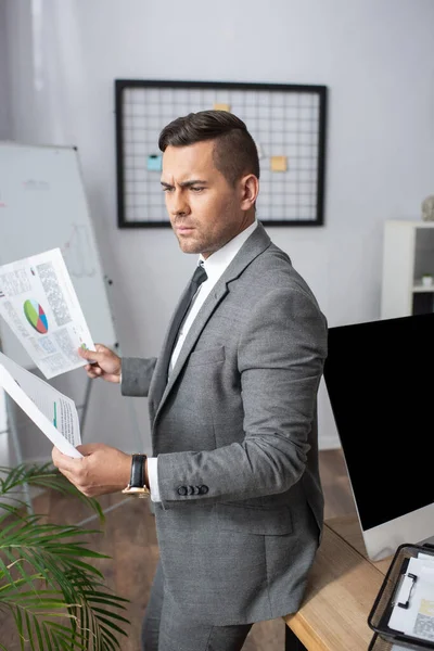 Comerciante pensativo em desgaste formal segurando infográficos no escritório — Fotografia de Stock