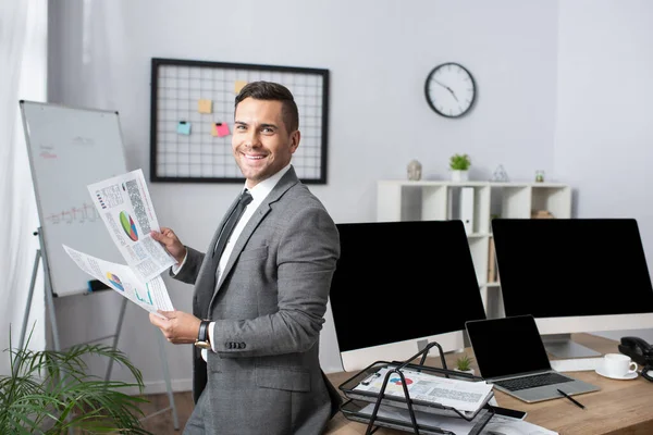 Commerciante sorridente che tiene infografica mentre in piedi vicino a monitor con schermo bianco — Foto stock