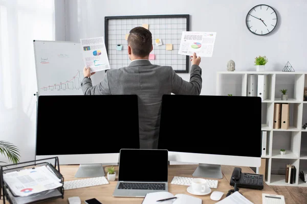 Back view of trader holding infographics near monitors with blank screen on blurred foreground — Stock Photo