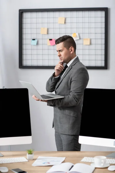 Comerciante reflexivo celebración de la computadora portátil, mientras que de pie en el lugar de trabajo cerca de monitores con pantalla en blanco en primer plano borrosa - foto de stock