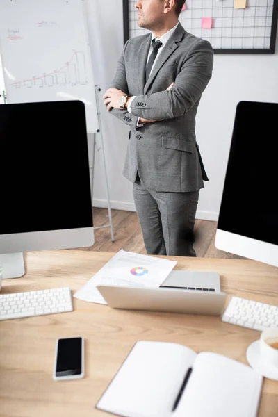 Vista parcial del comerciante de pie con brazos cruzados cerca de la computadora portátil y monitores con pantalla en blanco - foto de stock