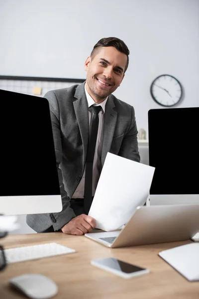 Smiling trader looking at camera near computer monitors and laptop on blurred foreground — Stock Photo