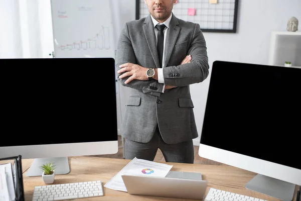 Cropped view of trader standing with crossed arms near laptop and computer monitors — Stock Photo