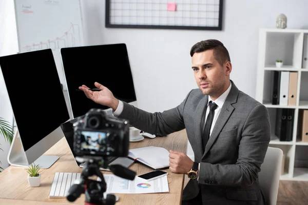 Trader pointing with hand at computer monitors during online streaming on digital camera, blurred foreground — Stock Photo