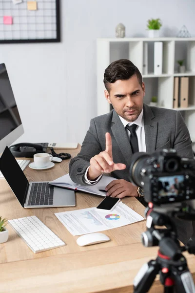 Trader pointing with finger near devices and charts during video recording on digital camera, blurred foreground — Stock Photo