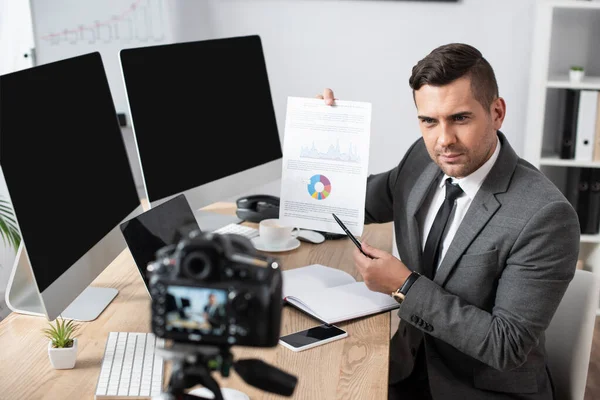 Comerciante apuntando con pluma a las infografías durante la grabación de vídeo en la cámara digital en primer plano borrosa - foto de stock