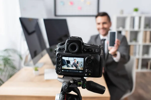 Foco seletivo da câmera digital perto de empresário segurando smartphone com tela em branco durante a transmissão de vídeo em fundo turvo — Fotografia de Stock