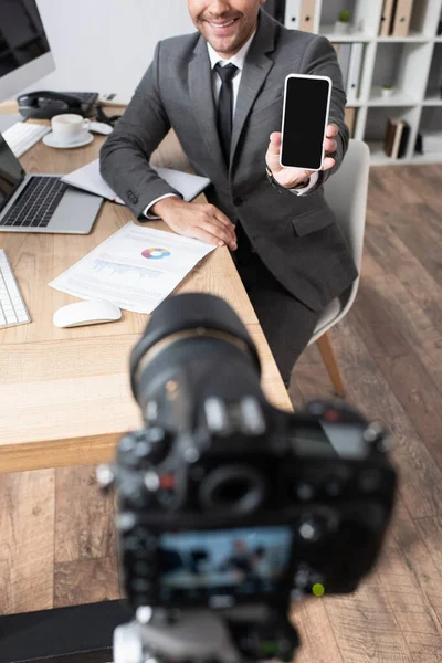 Vista ritagliata di uomo d'affari che mostra il telefono cellulare con schermo vuoto vicino alla fotocamera digitale in primo piano sfocato — Foto stock