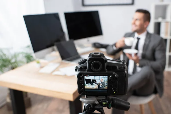 Selective focus of digital camera near businessman holding coffee cup while sitting near computers on blurred background — Stock Photo